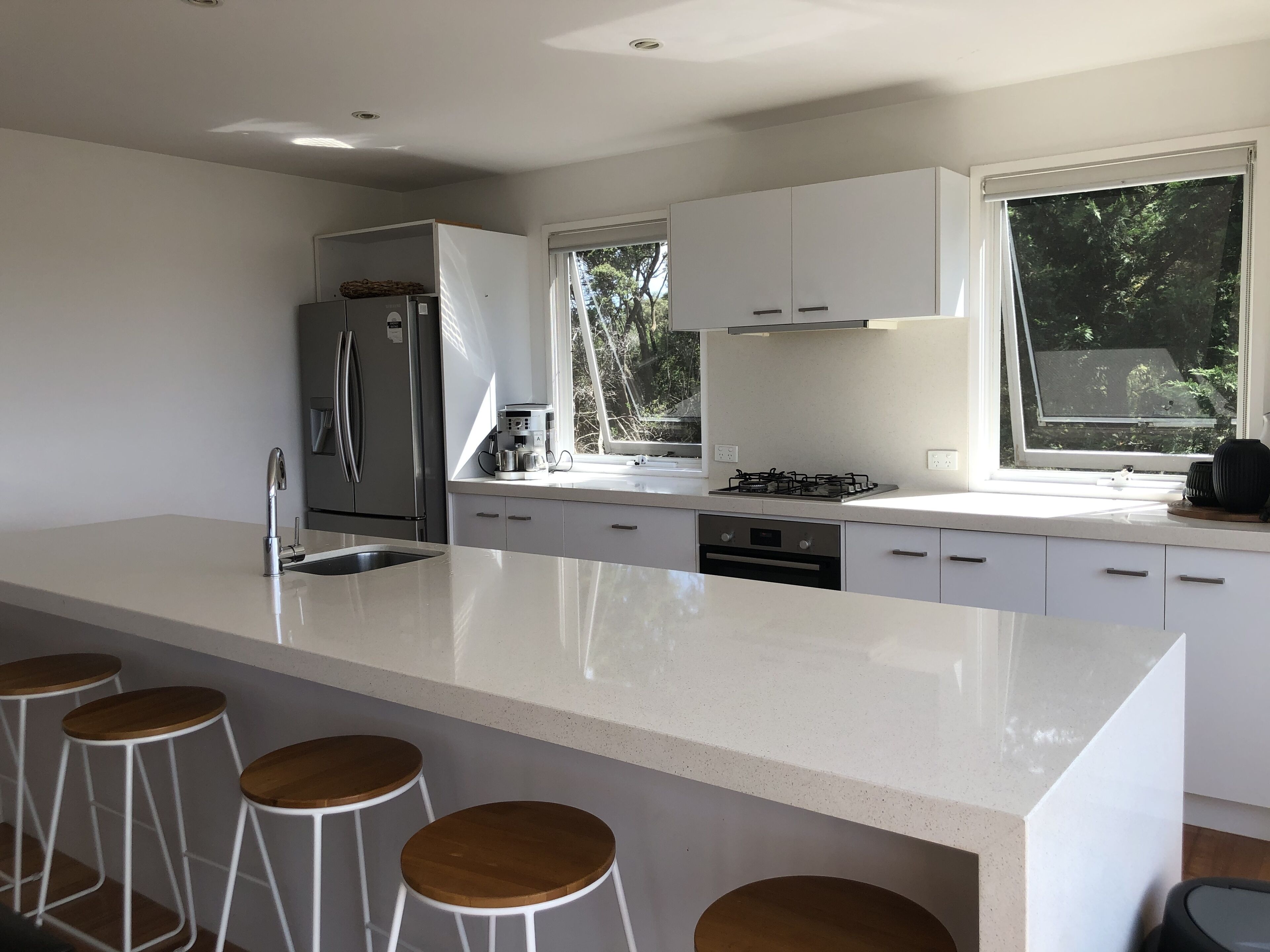 Een moderne keuken met witte kasten, een groot eiland met krukken, een inbouwoven en een raam met uitzicht op groen.