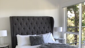 Een moderne slaapkamer met een groot bed, grijs en wit beddengoed, een zachte grijze plaid en uitzicht op bomen door een groot raam.