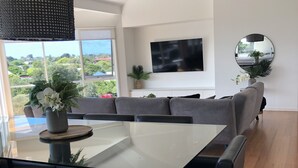Een moderne woonkamer met een glazen eettafel, zwarte stoelen, een grijze zithoek, een plafondventilator, een flatscreen-tv en een groot raam met uitzicht op groen en gebouwen.