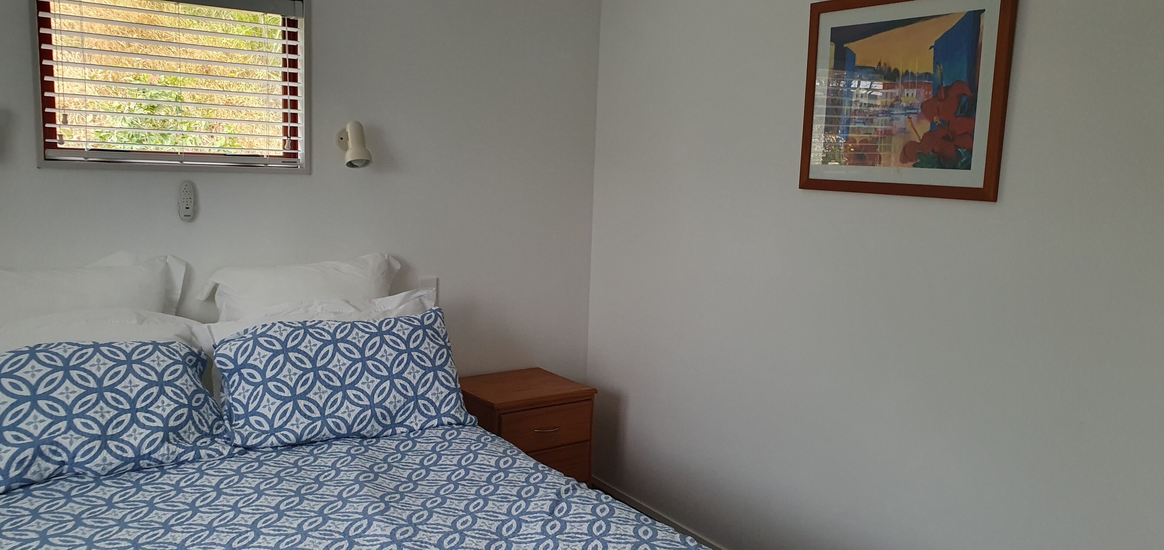 A bedroom with a bed, a nightstand, a wall-mounted light, a window with blinds, and a framed picture on the wall.
