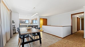 A modern kitchen with a central island, a dining area with a wooden table and chairs, and a view of the outside through large windows. - Grandview House - Ocean Views (KIANGA)