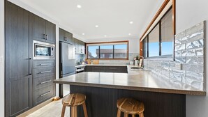 A modern kitchen with dark cabinetry, stainless steel appliances, a large island with stools, and a view through the window. - Grandview House - Ocean Views (KIANGA)