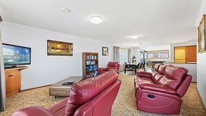 A spacious living room with leather recliners, a flat-screen TV, a bookshelf, and a dining area with a kitchen in the background. - Grandview House - Ocean Views (KIANGA)