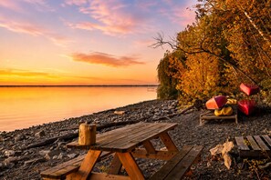 Ett picknickbord och några bänkar av trä står på en stenstrand, med höstlövsverk och en stilla sjö i bakgrunden.