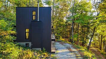 A modern, dark-colored building with a chimney, surrounded by a forested area with a gravel path leading to it.