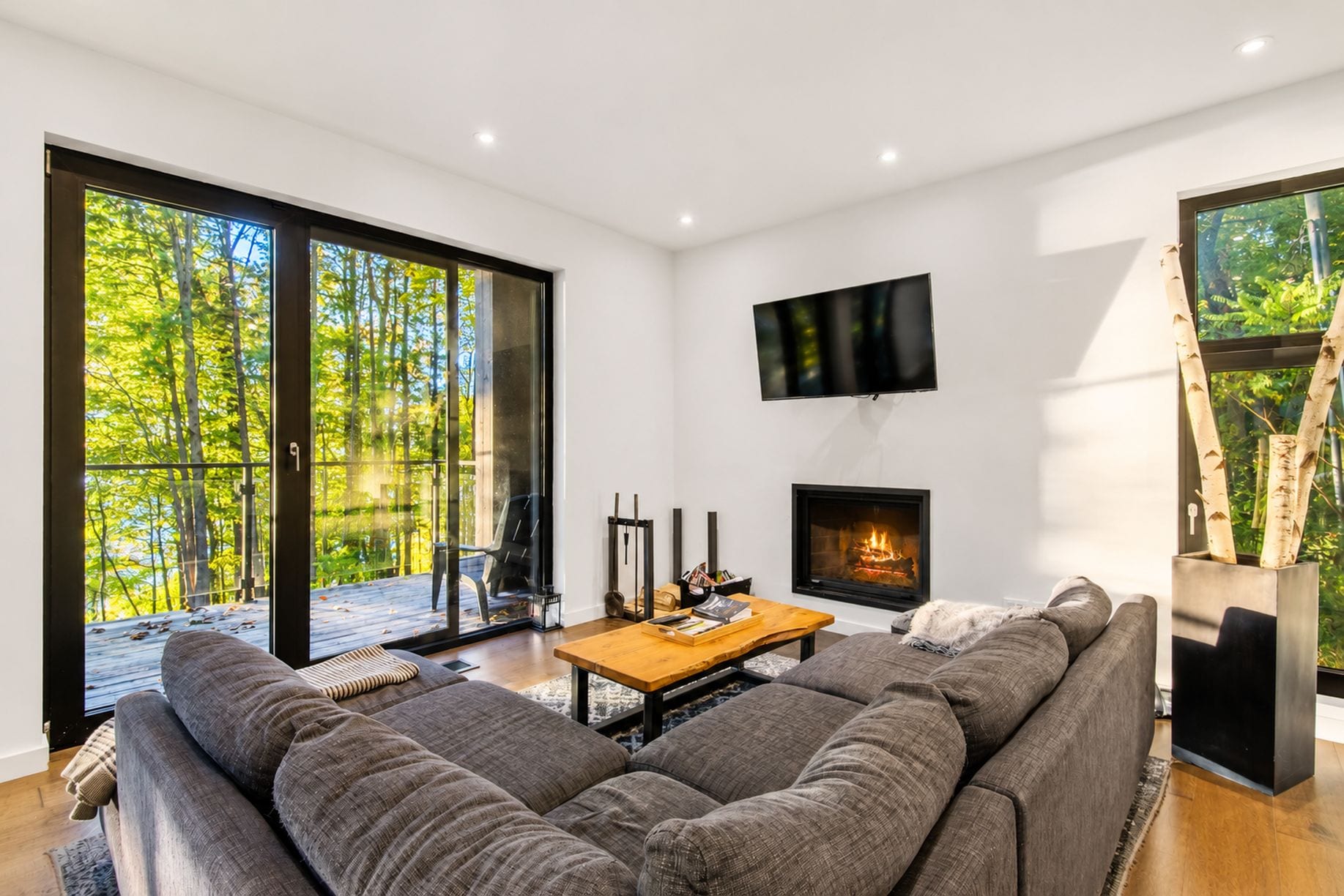 A modern living room with a large sofa, a fireplace, a television, and a sliding glass door leading to a deck.