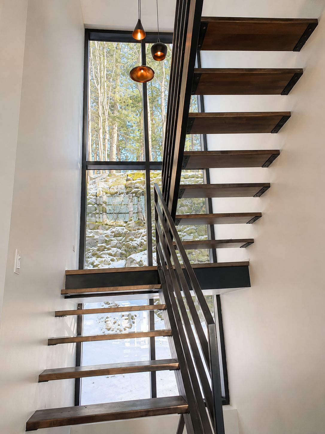 A modern staircase with a metal railing and wooden steps, leading to a large window with a view of trees and rocks.