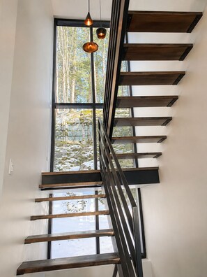 Una moderna scala con ringhiera in metallo e gradini in legno, che conduce a una grande finestra con vista su alberi e rocce.