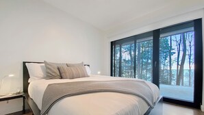 A modern bedroom with a large bed, a nightstand, a lamp, and a view of trees through a large window. - Cozy lakefront Retreat with Spa Pet Friendly (St-Armand)