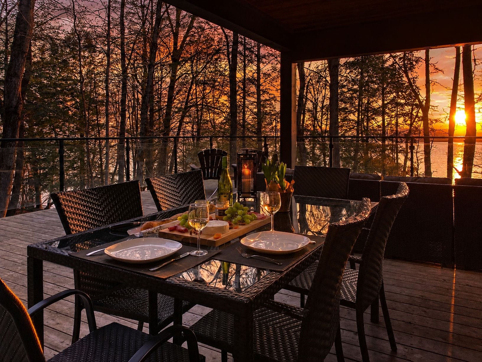 Un área de comedor al aire libre con una mesa preparada para dos personas, rodeada de sillas y con vista a un cuerpo de agua con árboles al fondo.