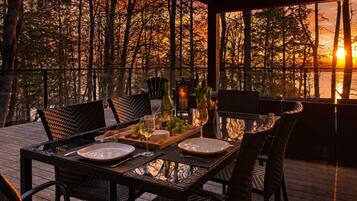 Un área de comedor al aire libre con una mesa preparada para dos personas, rodeada de sillas y con vista a un cuerpo de agua con árboles al fondo.