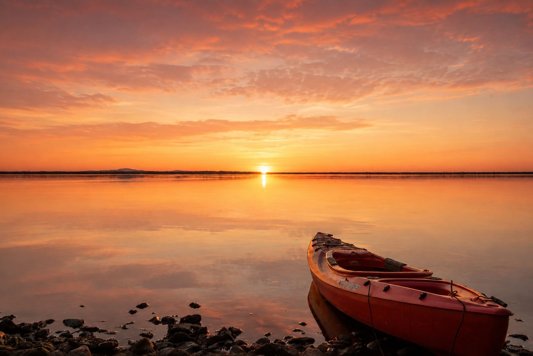 A calm body of water with a sunset, a rowboat, and a rocky shoreline.