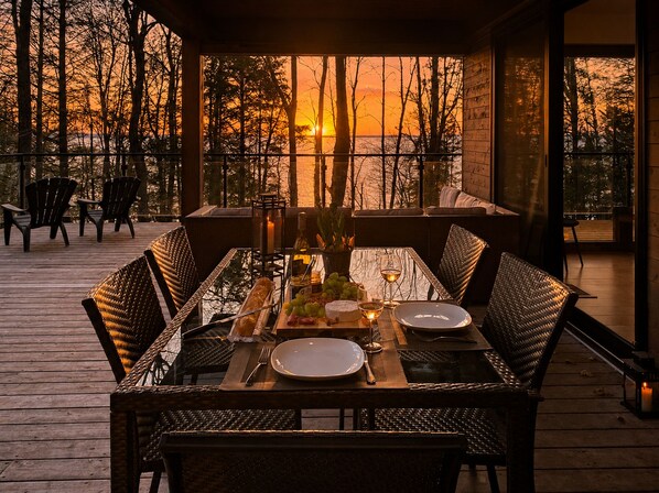 A dining area with a table set for two, surrounded by wicker chairs, overlooking a sunset view.