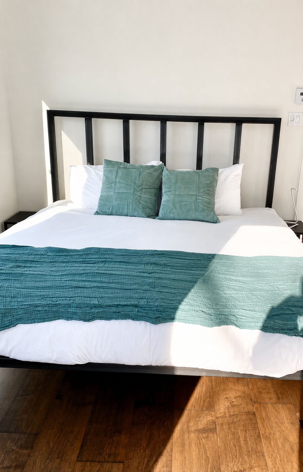 A bed with a white and teal bedspread, two pillows, and a black headboard in a room with wooden flooring.