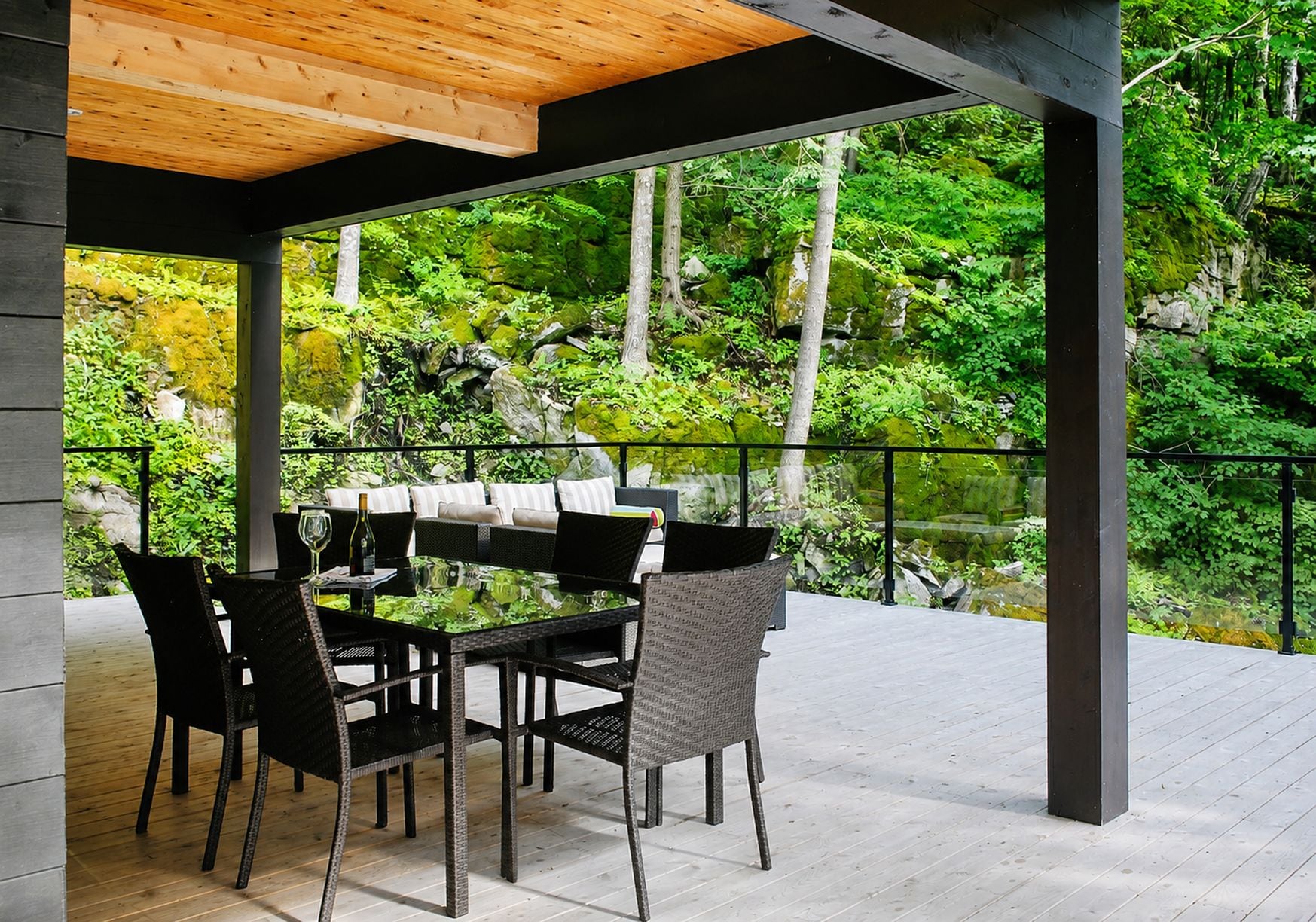 An outdoor patio with a dining table and chairs, surrounded by a wooden deck and a metal railing, overlooking a lush garden.