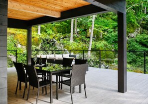 An outdoor patio with a dining table and chairs, surrounded by a wooden deck and a metal railing, overlooking a lush garden. - Lakefront Private Oasis Hot Tub Pet Friendly (St-Armand)