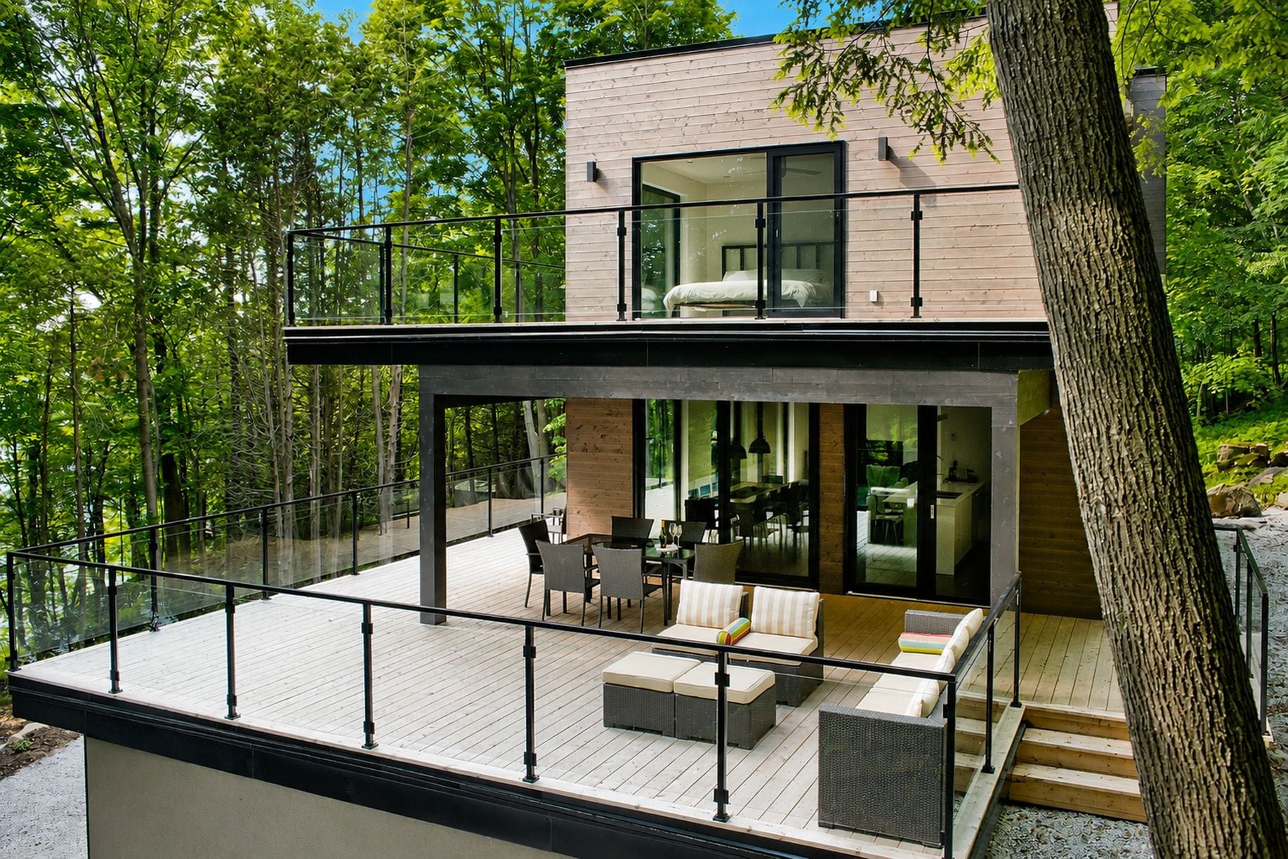A modern two-story house with a balcony, large windows, and a view of the surrounding forest.