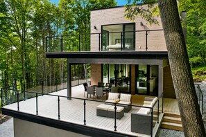 A modern two-story house with a balcony, large windows, and a view of the surrounding forest.