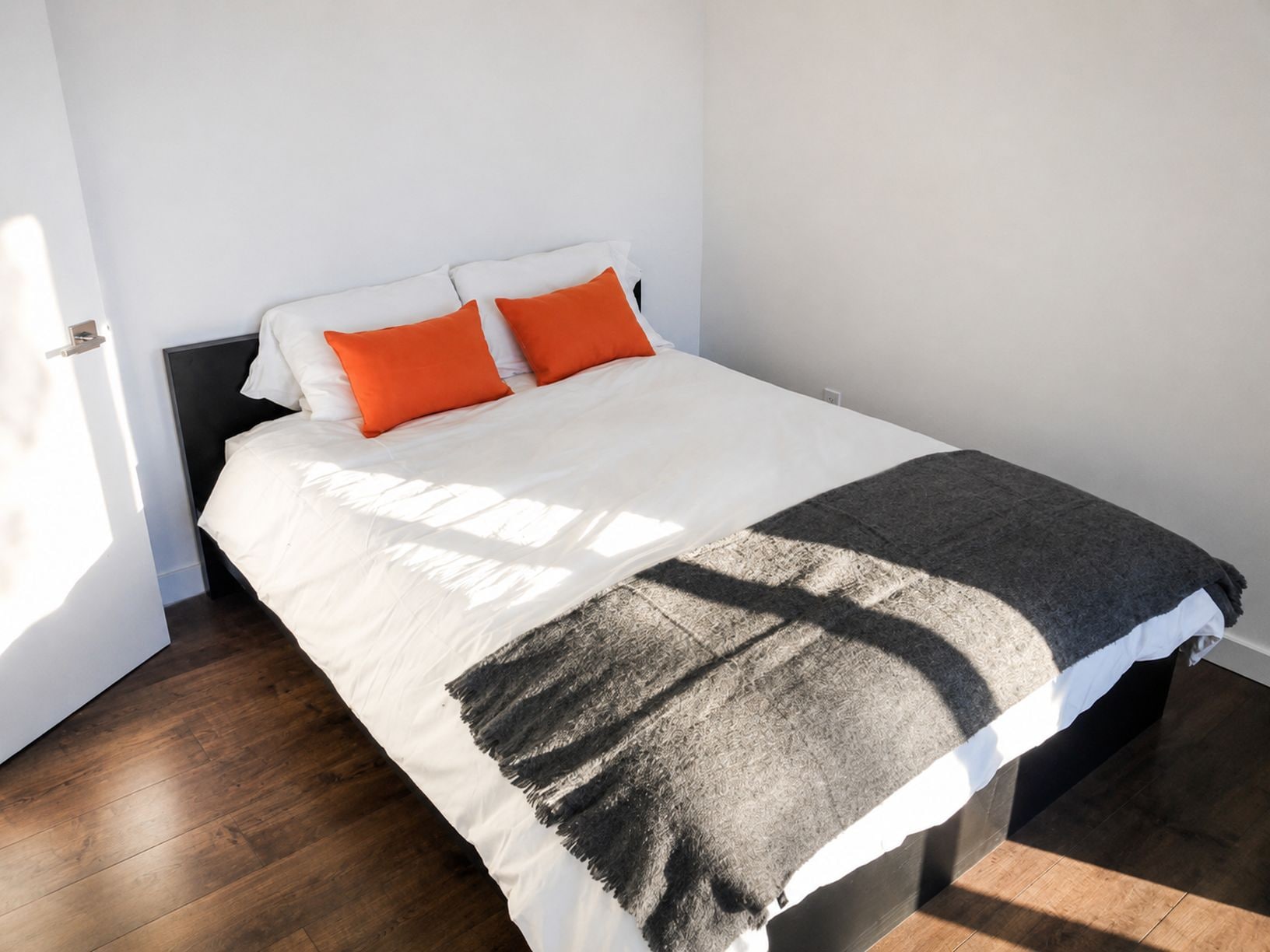 Una cama con ropa de cama blanca y dos almohadas naranjas, una manta gris y un cabecero oscuro.
