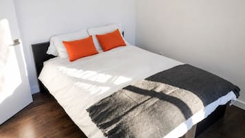 Una cama con ropa de cama blanca y dos almohadas naranjas, una manta gris y un cabecero oscuro.