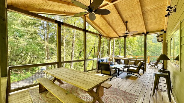 A wooden deck with a table and benches, surrounded by large windows offering a forest view. - Hosting a retreat, family reunion, girls or couples weekend or special occasion? (Ellijay)