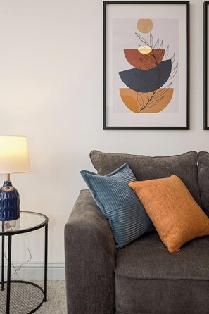 A living room with a grey sofa, a round side table with a lamp, and a framed abstract artwork on the wall.