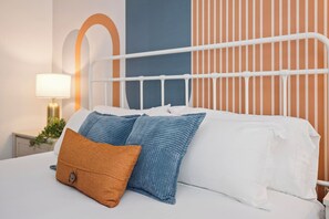 Cama bem arrumada com roupa de cama branca e duas almofadas, uma laranja e outra azul, em quarto com cabeceira de metal branca e parede listrada ao fundo.