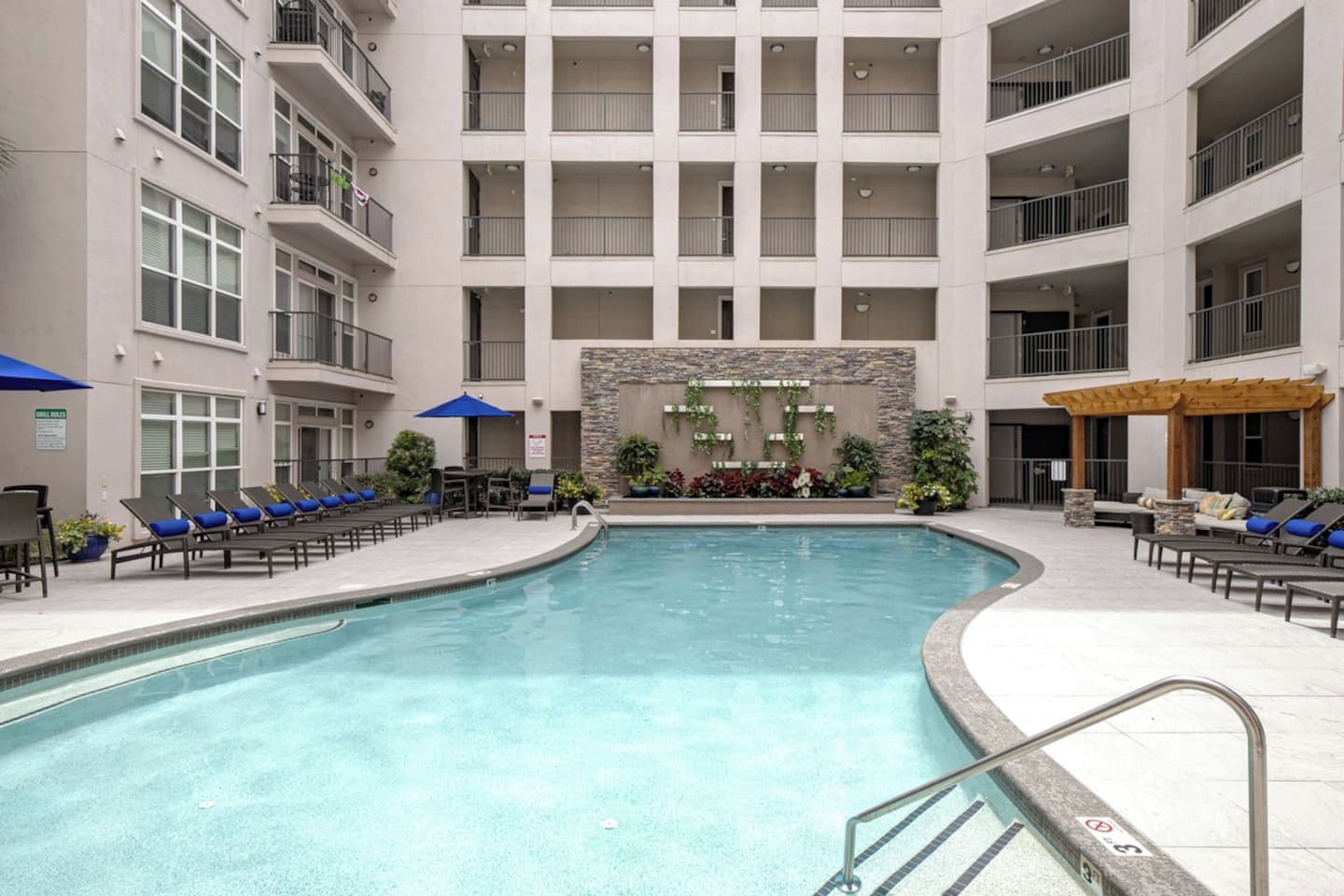 Área de piscina de hotel com várias espreguiçadeiras e guarda-sóis, uma parede de pedra com vegetação e um edifício com varandas.
