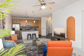 A modern living room with a sofa, coffee table, and a TV. There is a kitchen area with a refrigerator, microwave, and a dining space.