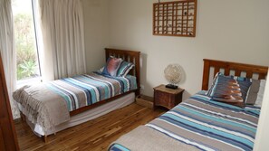 A bedroom with two beds, wooden flooring, a window with curtains, and a wall-mounted shelf with decorative items. - Driftaway -WIFI, NETFLIX, pet friendly, Comfort + Undercover deck with hammock (Marion Bay)
