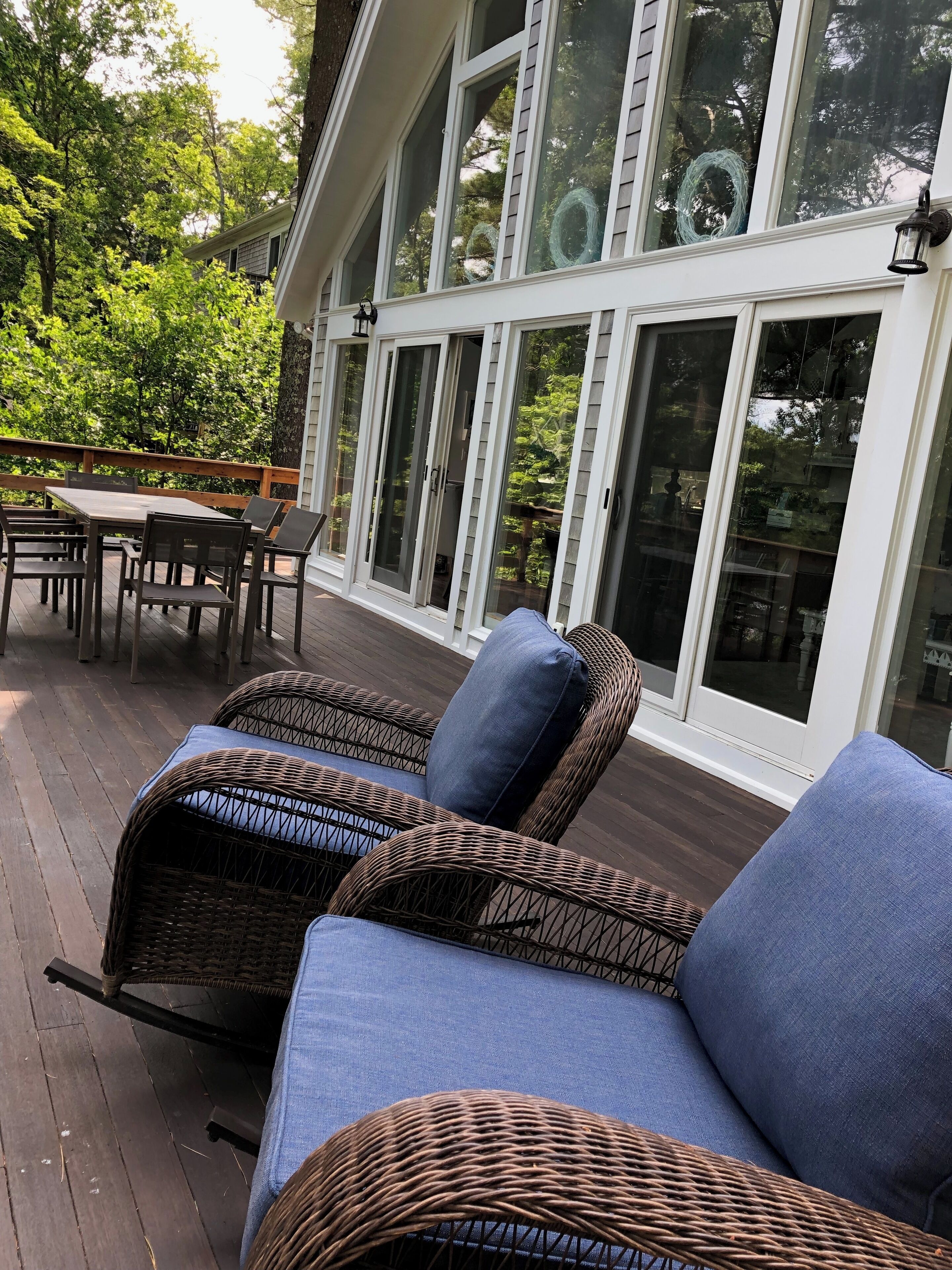 Une terrasse couverte, aménagée avec des meubles en osier et une table, de grandes portes vitrées et une vue sur les arbres.