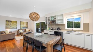 A modern kitchen with white cabinetry, a dining table with chairs, and a hanging light fixture. - Footsteps to the Bay by HolidayCo. (Pretty Beach)