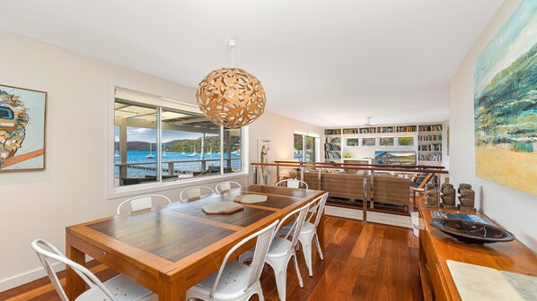 A spacious dining area with a wooden table and white chairs, overlooking a scenic harbor view through large windows. - Footsteps to the Bay by HolidayCo. (Pretty Beach)