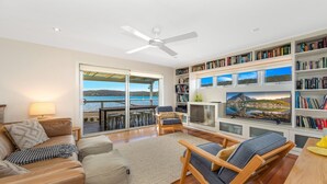 A spacious living room with a large bookshelf, a comfortable seating area, and a view of the sea through a sliding glass door. - Footsteps to the Bay by HolidayCo. (Pretty Beach)