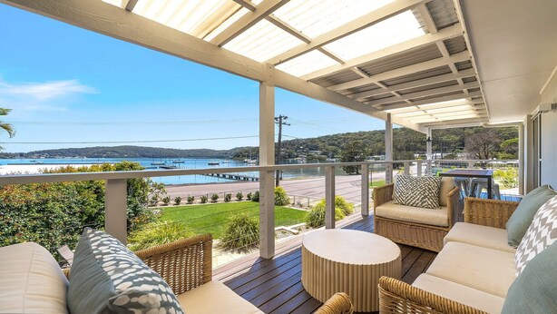 A covered outdoor seating area with wicker furniture, a round table, and a view of a lake with boats. - Footsteps to the Bay by HolidayCo. (Pretty Beach)