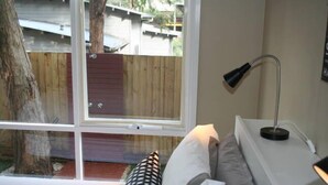 A bedroom with a large window, a bedside lamp, and a view of a wooden deck and trees.