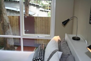 A bedroom with a large window, a bedside lamp, and a view of a wooden deck and trees. - Lorne Surfdogz at Lorne (Lorne)