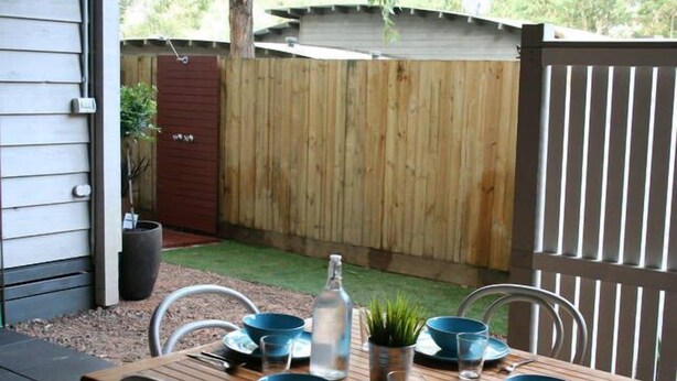 A wooden table set with blue bowls, plates, and a bottle, surrounded by metal chairs, in a backyard with a wooden fence and a small garden area.