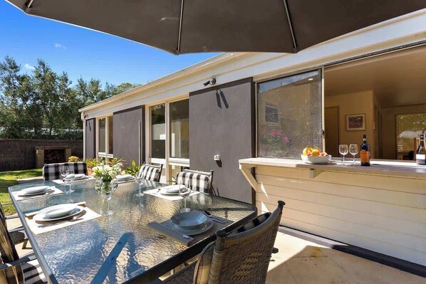 An outdoor dining area with a glass table, metal chairs, and a large umbrella.