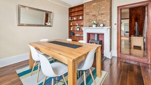 A dining area with a wooden table and white chairs, a brick fireplace, and a bookshelf.