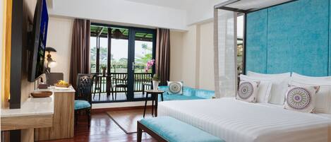 A hotel room with a bed, a desk, a TV, a balcony with chairs and a table, and a view of greenery.