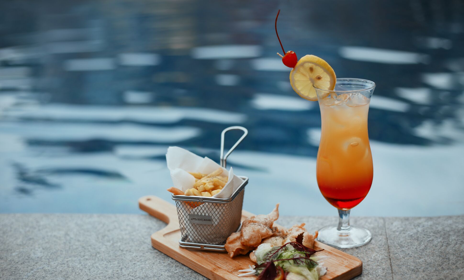 A glass of red cocktail with a lemon slice and cherry garnish, a basket of fries, and a salad on a wooden board.