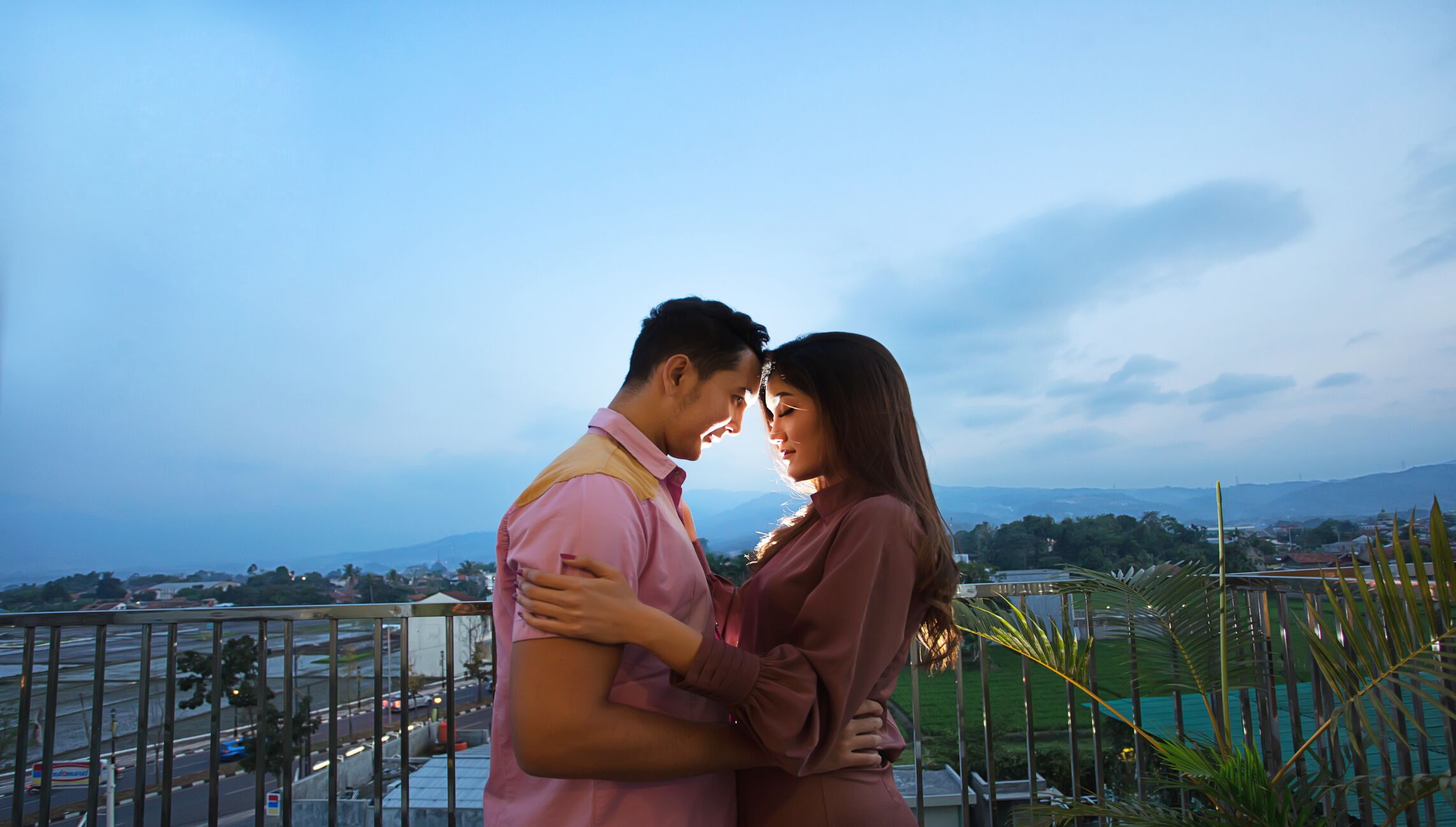 A couple embracing on a balcony overlooking a cityscape with distant mountains.