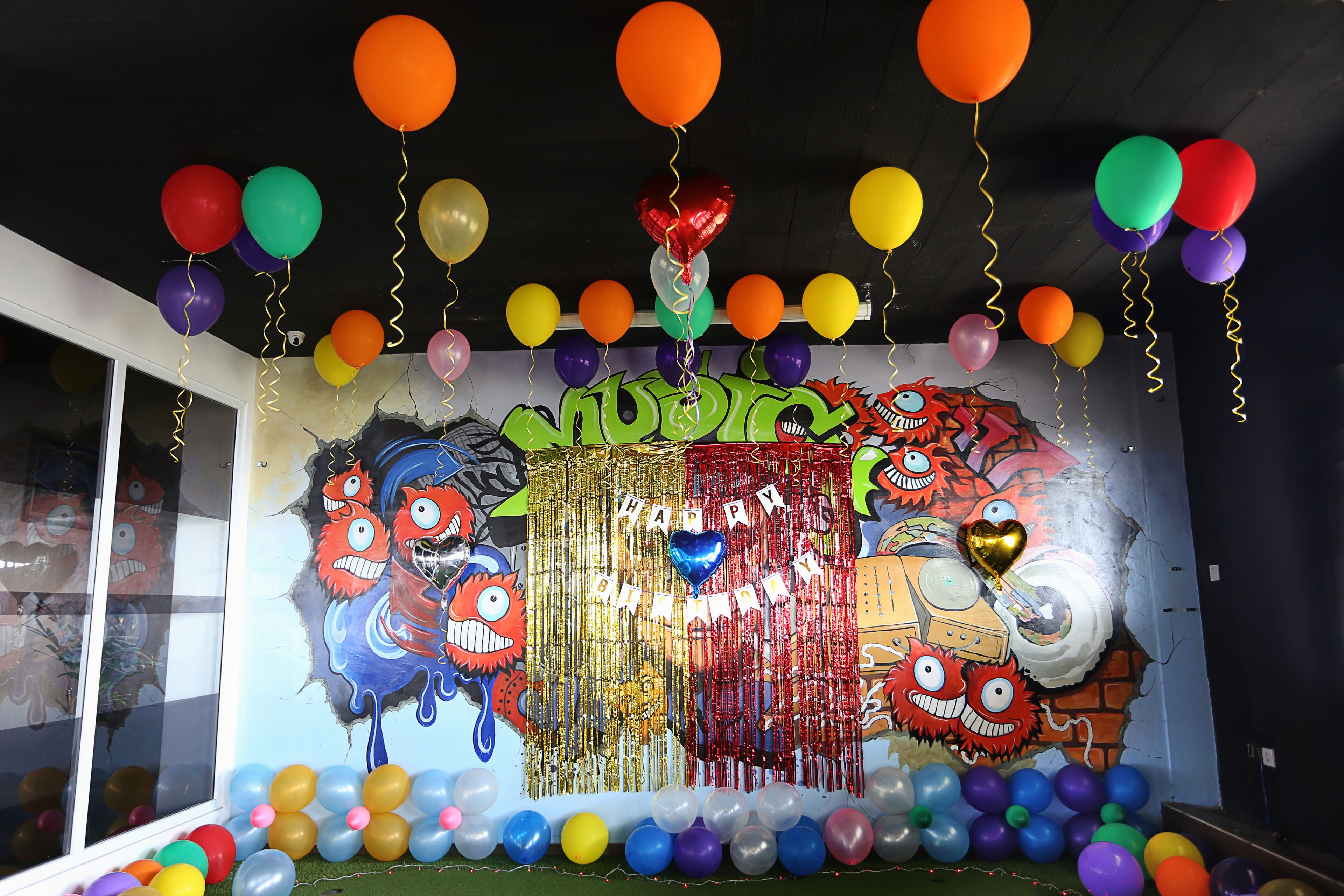 A room with a colorful balloon arrangement and a birthday-themed mural on the wall.