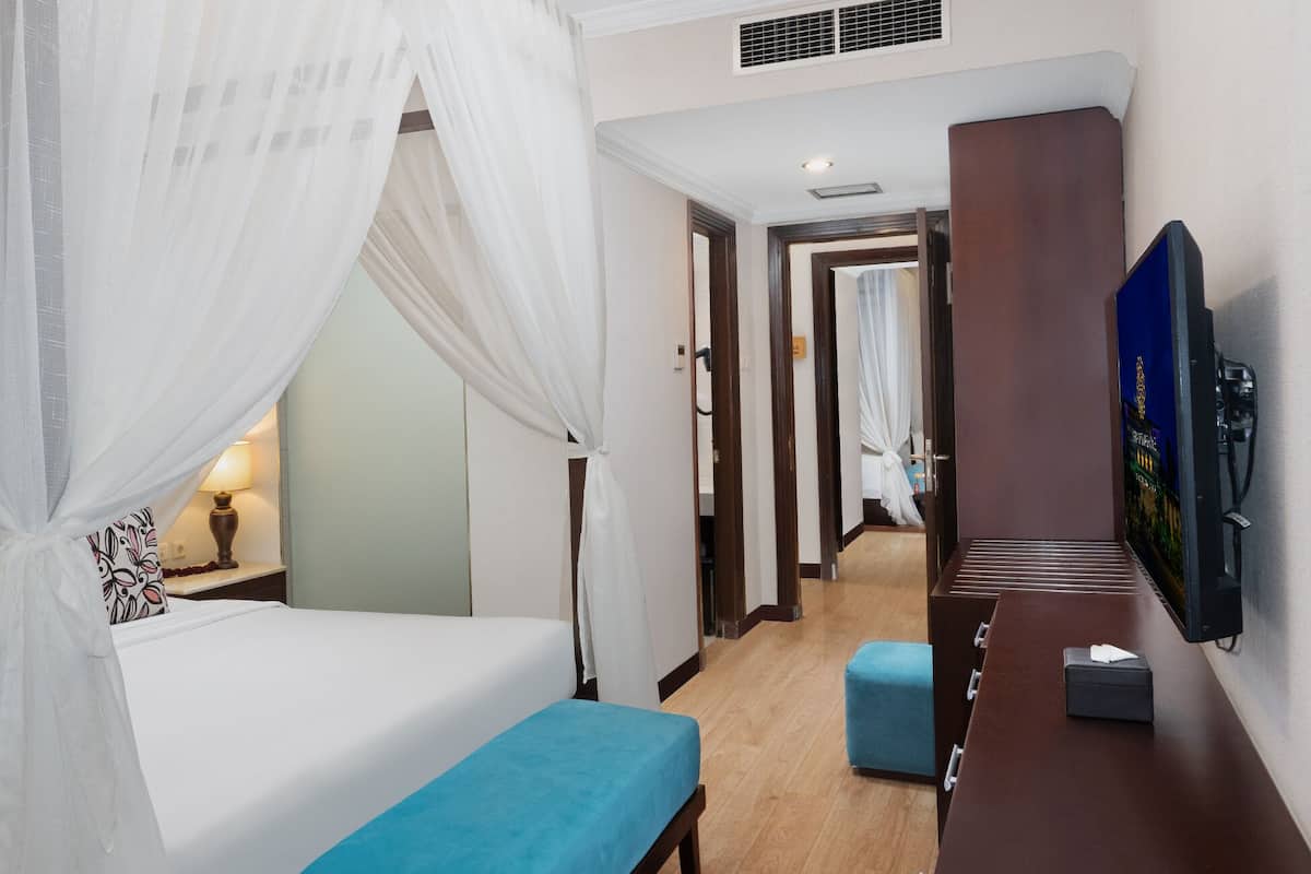 Une chambre d’hôtel avec un lit, une table de chevet, une lampe, un banc, une télévision et un couloir menant à une autre pièce.