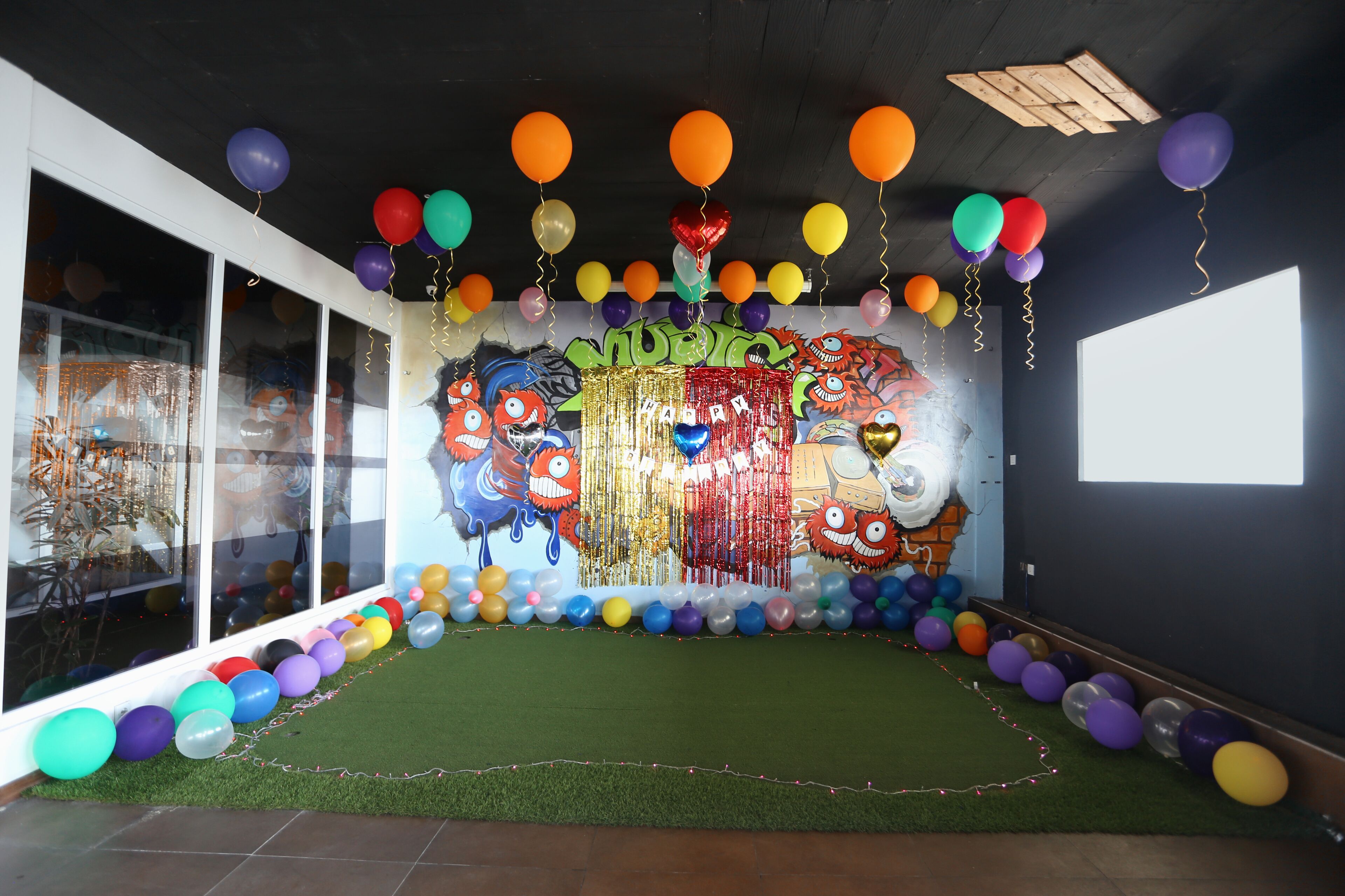 A room with a green artificial turf floor, large windows, and a wall decorated with colorful balloons and a cartoon character mural.