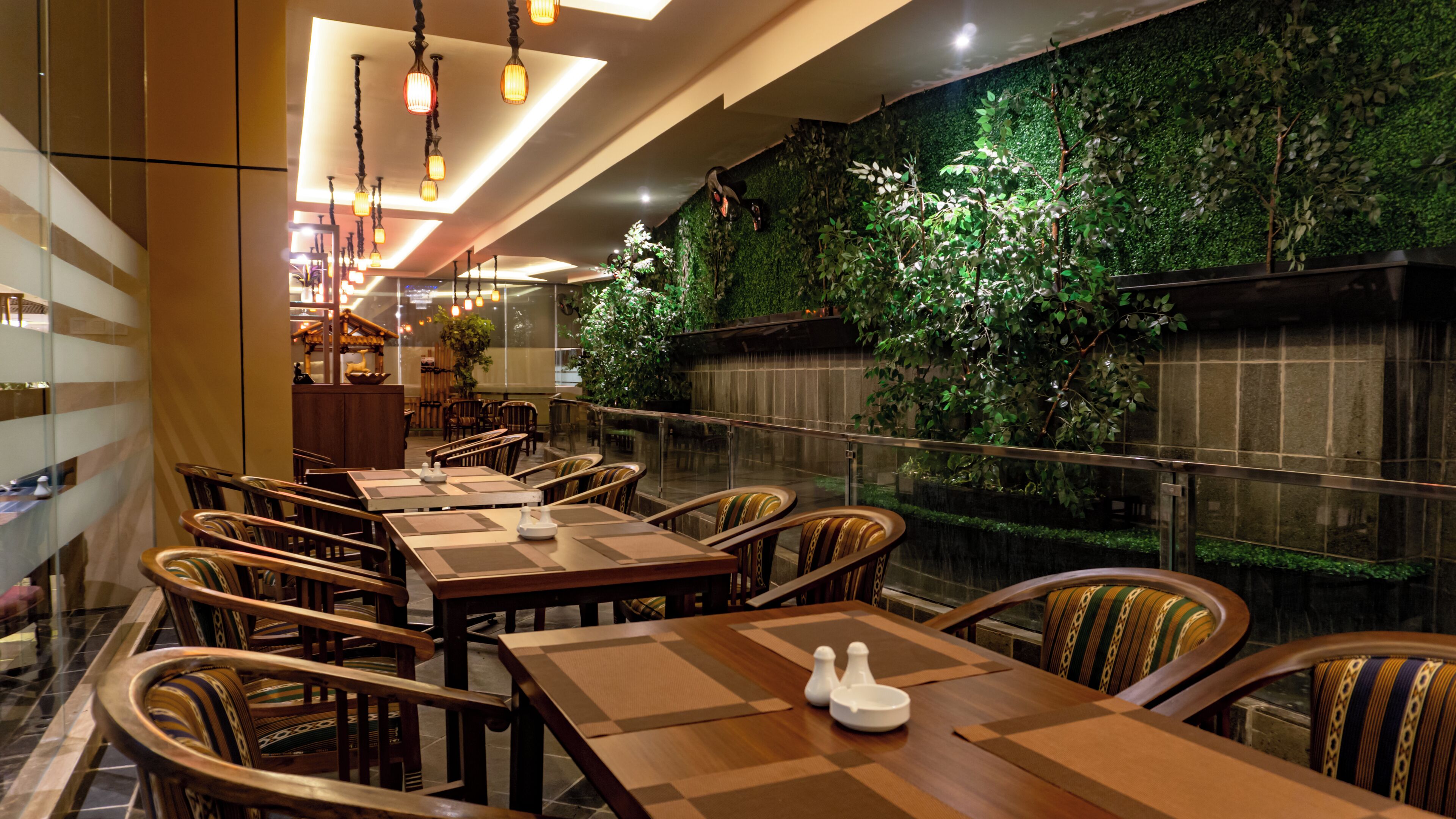 A restaurant with wooden tables and chairs, a green wall with plants, and hanging lights.