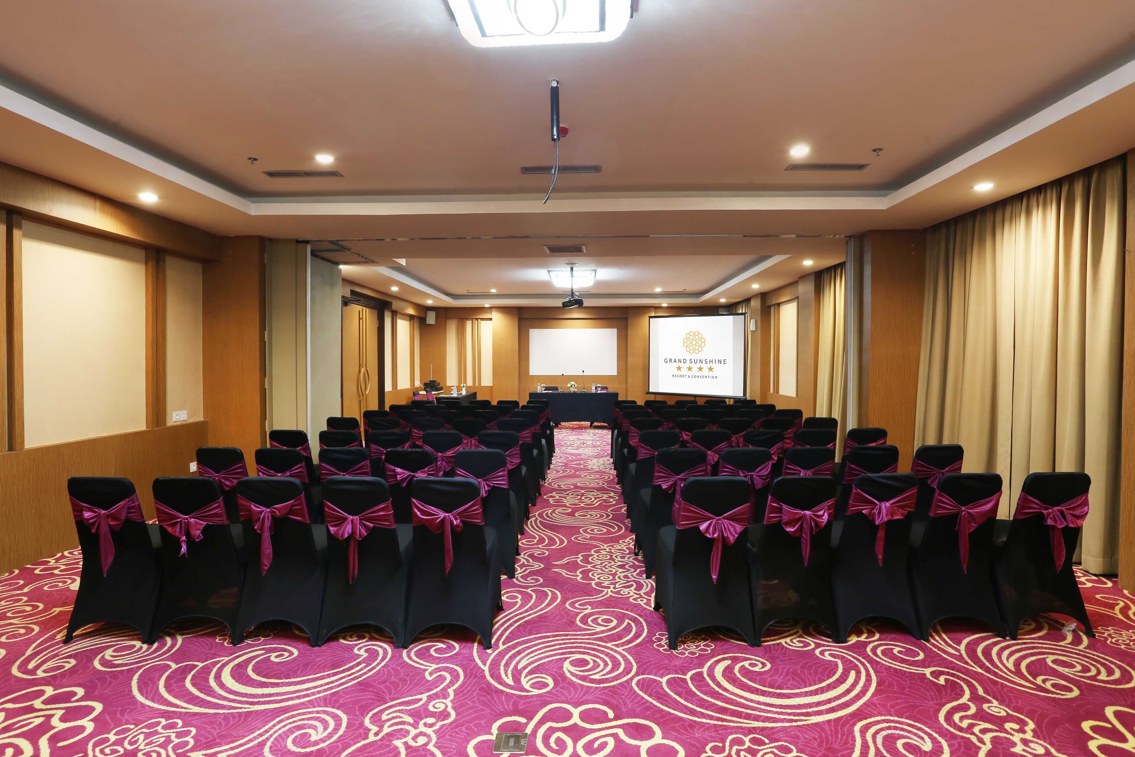 A conference room with rows of black chairs with pink sashes, a large screen at the front, and a red carpet with a swirl pattern.