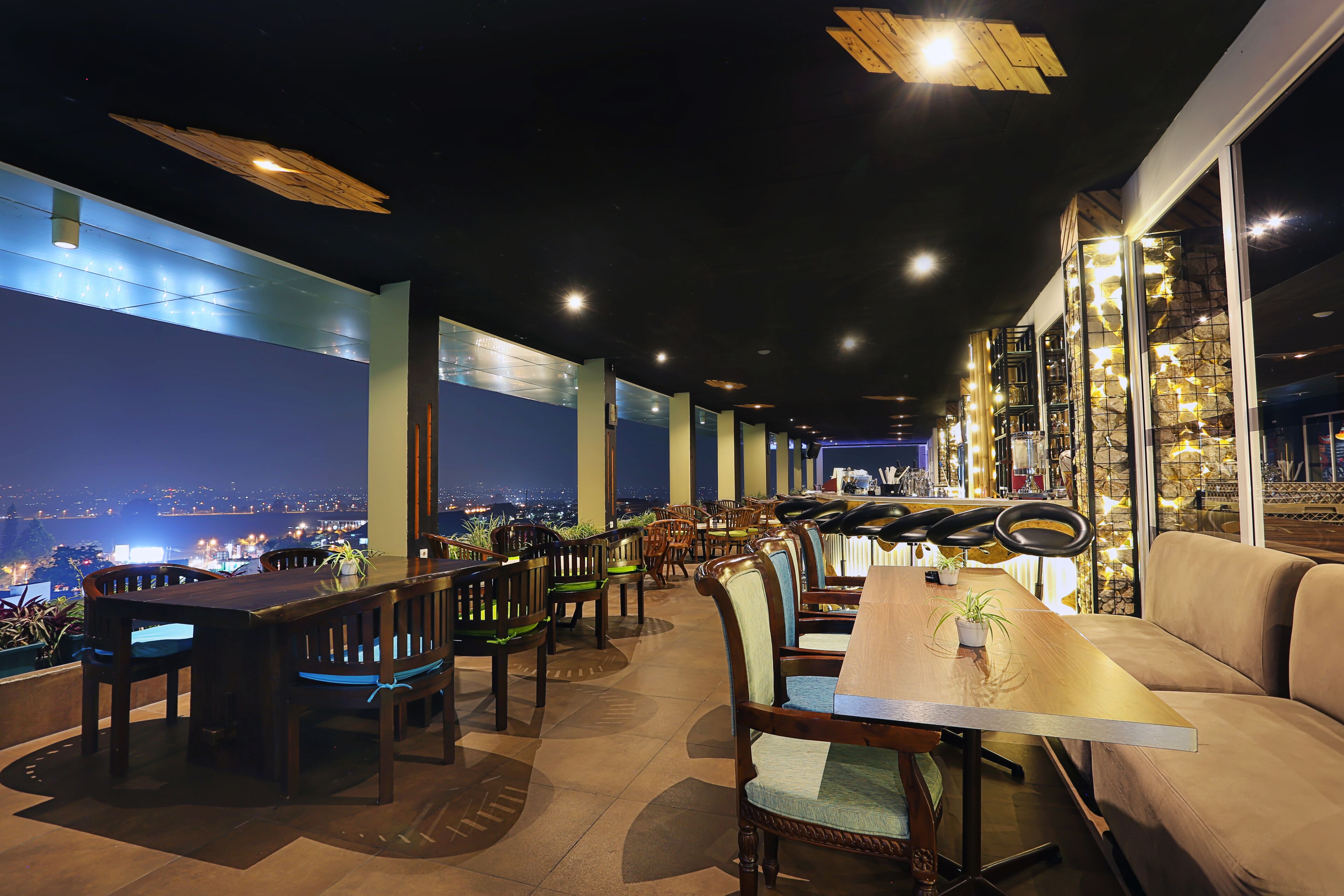Un comedor interior con mesas y sillas, ventanas amplias y vistas de la ciudad de noche.