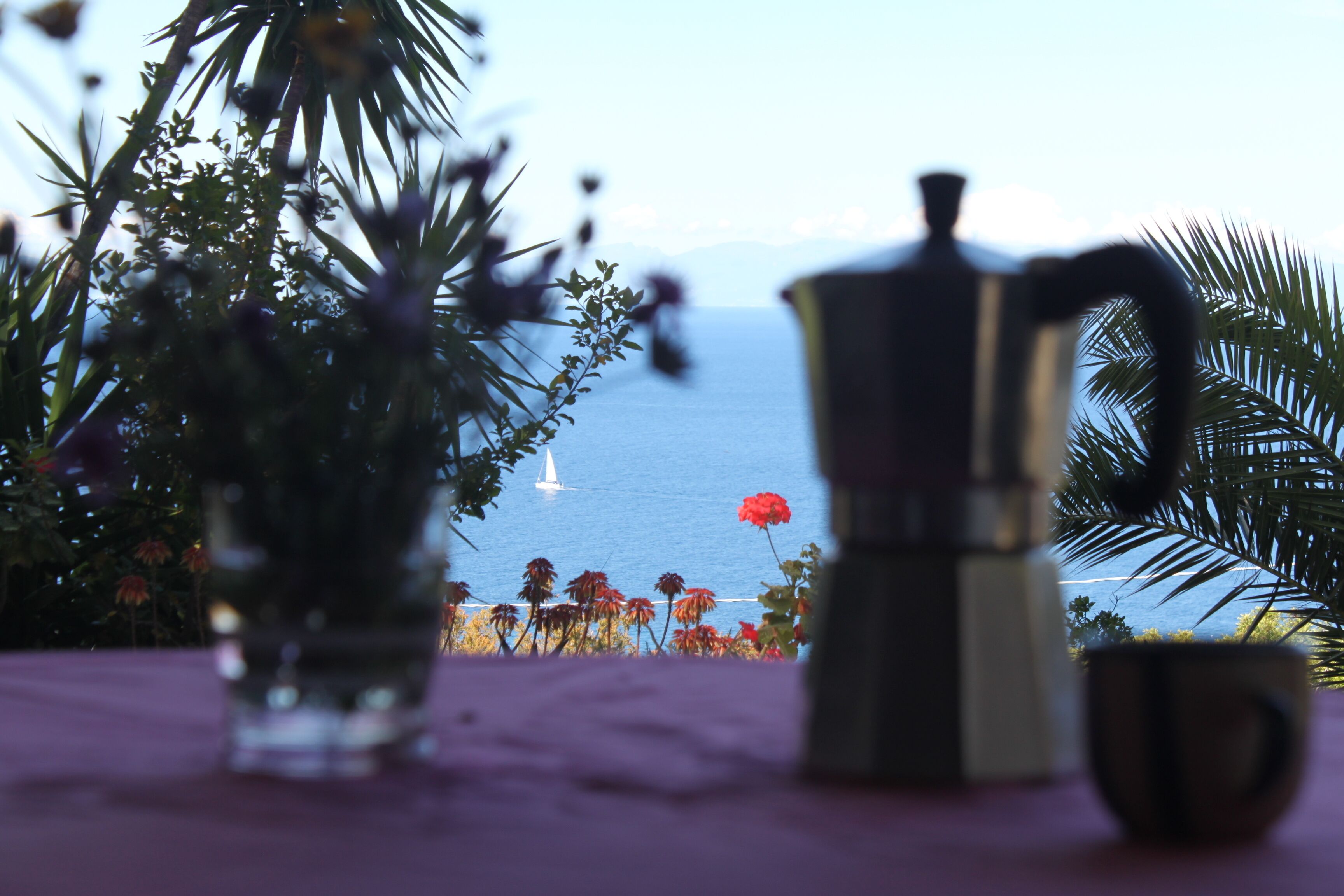Ein Tisch mit einer Kaffeemaschine und einer Tasse, mit Blick auf ein Gewässer und ein Segelboot.
